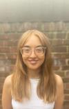 Lillian Race smiling against a brick wall. She has mid-length strawberry blonde hair. She wears glasses and is wearing a white sleeveless top.