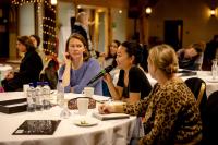 Three woman sat on a table at a work event. There are items scattered on the table including four bottles of water, mugs, a notebook, a pencil case and a small plate. Two white women are looking at an Asian woman holding a microphone (presumably speaking through it). The background is blurred, but there are other tables and guests.