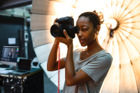 a Black woman with short black hair wearing a grey t-shirt takes a photo with an industrial camera. she is stood in front of a photographer's umbrella with a computer to her left