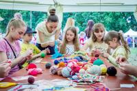 Two women help a group of children with a creative project. They are working as volunteers, explaining to the children how to knit with colourful wool