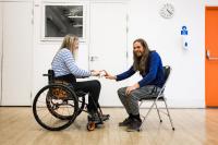 Image shows two people seated, one in a wheelchair, touching hands