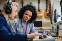 A music teacher helps a student during a music lesson. The student is playing a keyboard and the teacher is watching on and smiling