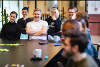 People sat together around a black table in an office. They are smiling and laughing. On the table, there is a mug, post-it notes, and a notebook.