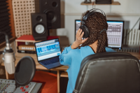 A young person sits at a music production desk. She is holding headphones with her back to the camera