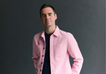 Joshua McTaggart stands in front of a grey background wearing a pink jacket