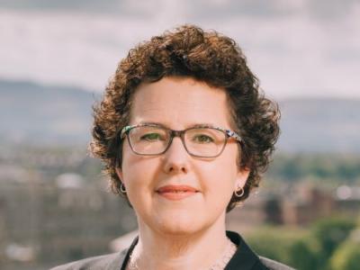 Joanna Baker has short, brown curly hair and wears glasses, small hoop earrings and nude lipstick. She is photographed in front of a viewpoint. The background is blurred.