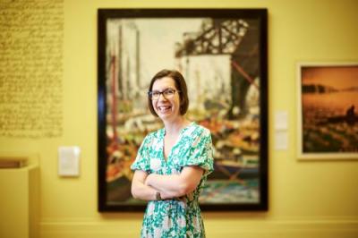 Dr Inbal Livne, Manchester Art Gallery Senior Creative Lead. She stands with her arms folded in front of a painting in an exhibition. She wears a green dress with white flowers, has short brown hair and wears black glasses. She is smiling and looking at the camera.