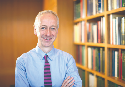 Roly Keating, a white man with grey hair smiles with his arms folded in front of bookshelves