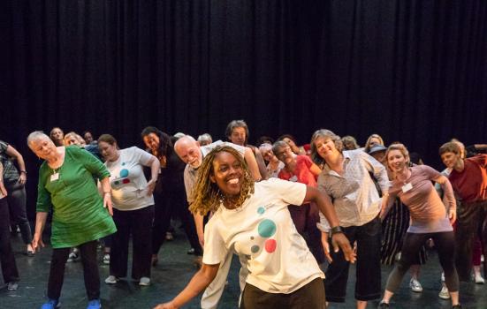 A group of community dancers of different ages on a stage in front of a black curtain