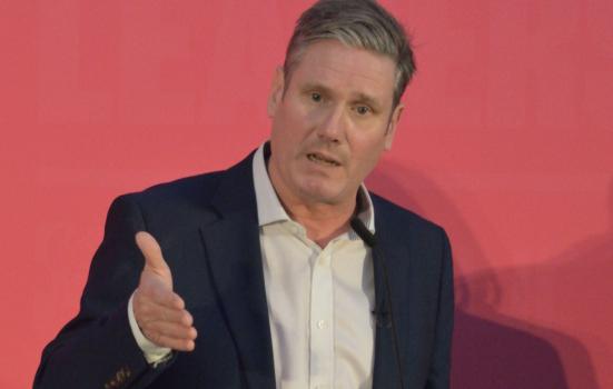 Keir Starmer speaking at a press conference. He is standing in front of a plan red background and wearing a red suit