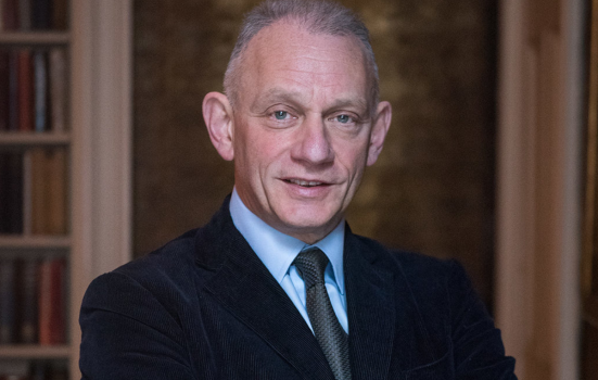 Portrait of Neil Mendoza. he is looking at the camera with his arms folded, wearing a black suit, blue shirt and tie