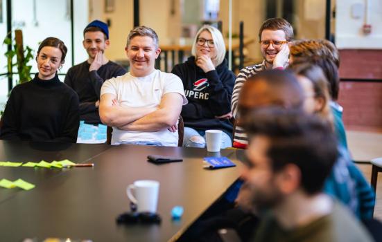 People sat together around a black table in an office. They are smiling and laughing. On the table, there is a mug, post-it notes, and a notebook.