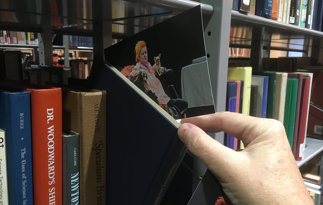 Photo showing a postcard being placed in a book on a bookshelf