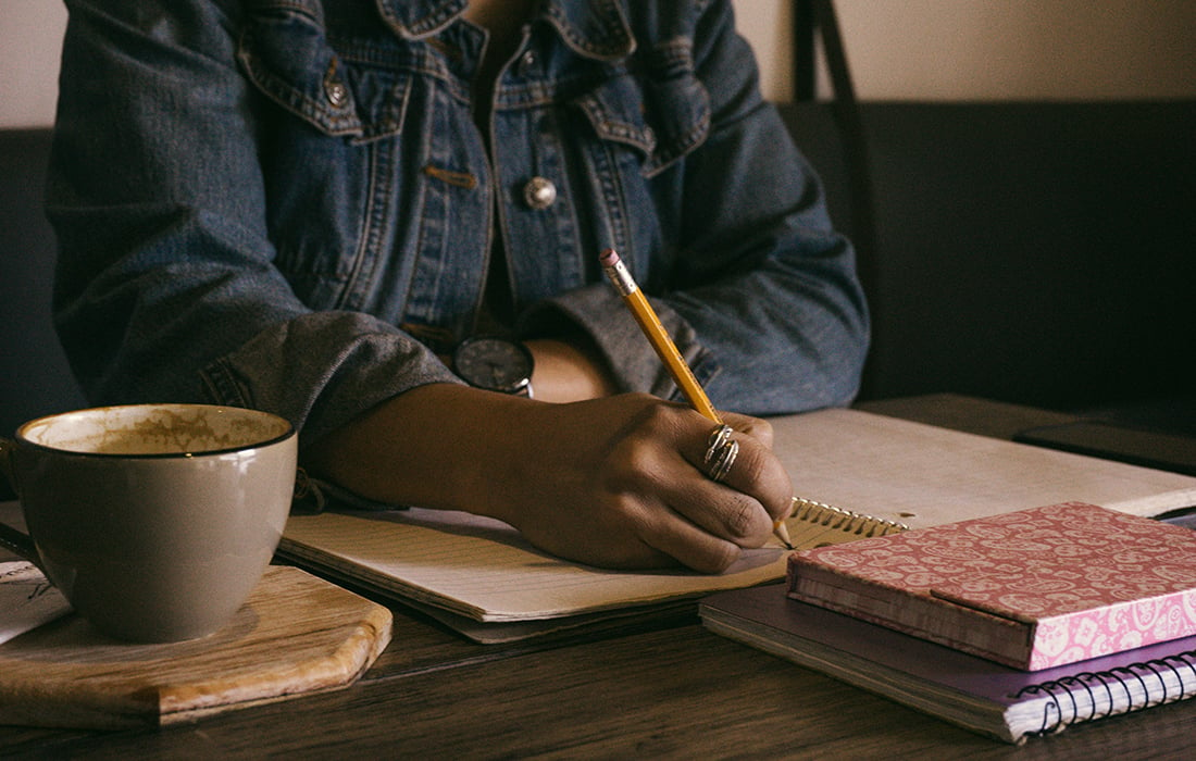 Black female sitting at a desk, writing with a pencil. Coffee cup and saucer in the foreground