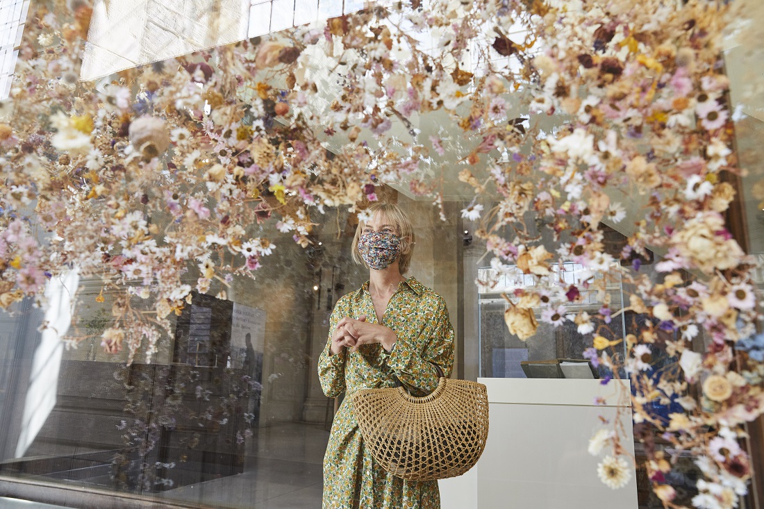 A woman in a floral dress and face mask looking an art installing of floating flowers