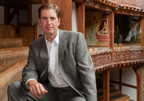 Neil Constable is sat inside Shakespeare's Globe. He has dark brown hair and is wearing a grey suit.