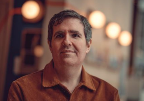 Headshot of David Byrne. He wears an orange, corduroy shirt. He has grey hair, bushy eyebrows and is slightly smiling, looking upwards of the camera.