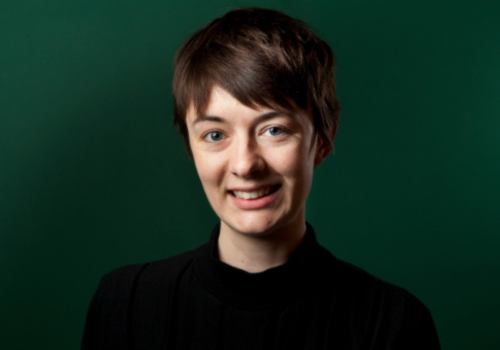 Lauren Clancy. She is smiling against a dark green wall. She is wearing a black top and has short brown hair.