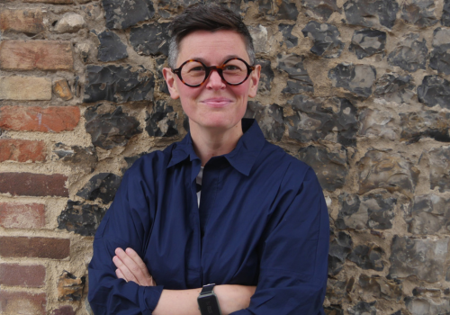 Peggy Hughes. She is photographed against a brick wall. She has short hair and wears round glasses.