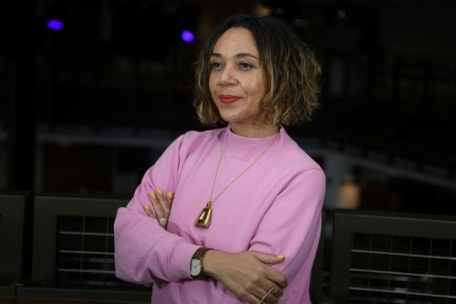 A photo of Raidene, a brown-skinned female, aged early forties, with wavy brown shoulder-length hair, wearing a pink jumper, looking away from the camera, smiling